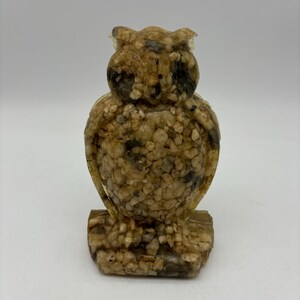 May include: A small owl figurine made of resin and embedded with small stones. The owl is brown and beige with a light yellow tint.