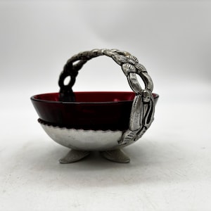 May include: A small bowl featuring a deep red glass insert held within a silver-toned metal base. The base has three short feet and a decorative arched handle adorned with a repeating floral pattern and textured metal detailing throughout.