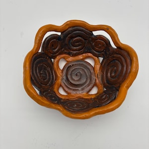 May include: A handmade ceramic bowl with a unique design. The bowl features a spiral pattern in shades of brown and orange. The outer rim is a vibrant orange, while the interior spirals are a darker brown. The bowl is approximately 6 inches in diameter.