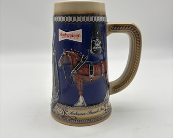 May include: A ceramic beer stein with a blue background and a brown horse illustration. The stein has a tan handle and rim, with the Budweiser logo and "Anheuser-Busch Inc" text. The stein is approximately 6 inches tall.