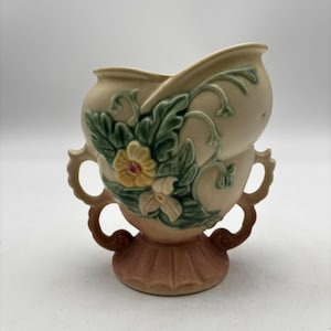 May include: A cream-colored ceramic vase featuring a raised relief design of green leaves and yellow flowers. The vessel has two ornate, curved handles on the sides and a fluted, pedestal-style base with a soft pinkish-brown hue at the bottom.