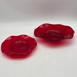 May include: Two red glass bowls with wavy edges. The bowls have a glossy finish and a dark rim. One bowl is smaller and flatter than the other, which is deeper. The bowls are likely decorative or for serving.