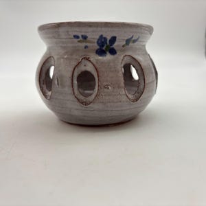 May include: A handcrafted, ceramic candle holder with a light gray finish. The holder features several oval cutouts and is adorned with blue floral accents. The rim and cutouts have a darker, reddish-brown hue, adding contrast to the design.