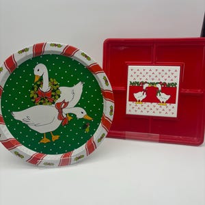 May include: Two Christmas themed metal trays. One is round with a green background and white geese with red bows. The other is square with a red background and a white goose with a red bow.
