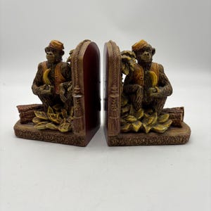 May include: A pair of decorative bookends featuring monkey figures. Each monkey is seated, holding a banana, and wearing a vest and hat. The bookends have a brown and gold color scheme with leaf and log details.