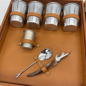 May include: A brown leather travel case with a set of six silver cups, a small silver cup, a silver spoon, and a bottle opener. The cups have brown leather bands around them.
