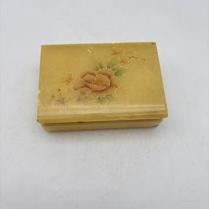 May include: A rectangular, yellow jewelry box with a floral design. The box features a large, peach-colored flower with green leaves and smaller orange flowers. The box is made of a smooth material and has a hinged lid.