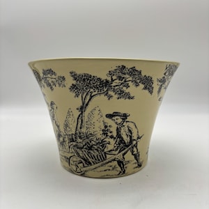May include: Cream-colored ceramic planter with a black toile design. The illustration depicts a man pushing a wheelbarrow, trees, and other figures in a pastoral scene. The planter has a flared shape.