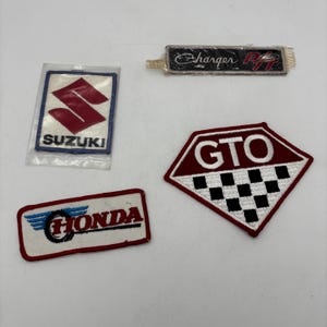 May include: Four embroidered patches featuring automotive branding. One rectangular Suzuki patch in plastic, one rectangular Charger R/T patch, one rectangular Honda patch with a winged tire logo, and one pentagonal GTO patch with a checkered pattern.