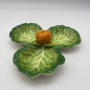 May include: A ceramic serving dish in the shape of three overlapping leaves, painted in shades of green and yellow. An orange fruit sits in the center. The dish is likely used for serving snacks or small portions.