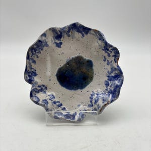 May include: A handmade ceramic dish with a unique, organic shape. The dish features a white base with blue splatter and a dark blue center. The rim has a wavy, irregular edge, and the overall design is artistic and rustic.