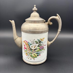 May include: A vintage white porcelain teapot with a silver-toned lid, handle, and spout. The teapot is decorated with a floral design featuring calla lilies and other colorful flowers. The handle has a curved design.