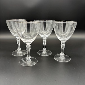 May include: Four clear stemmed glasses with a silver rim. The glasses have a wide bowl and a detailed stem. The glasses are arranged in a row on a dark surface.
