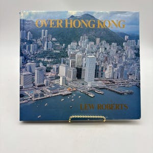 May include: A book titled "Over Hong Kong" by Lew Roberts. The cover features an aerial view of Hong Kong with skyscrapers, a harbor, and a mountain in the background. The text is in gold.