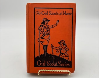 Könnte beinhalten: Ein orangefarbenes Buch mit dem Titel "The Girl Scouts at Home" mit schwarzen Illustrationen von Pfadfinderinnen. Das Buch gehört zur "Girl Scout Series". Das Buch wird von einem goldfarbenen Ständer gehalten.
