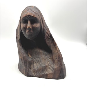 May include: A dark brown wooden sculpture carved into the shape of a human head and shoulders, featuring a draped hood-like covering over the head. The wood grain is visible throughout the surface, showing natural textures and carved contours.