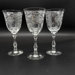May include: Three clear wine glasses with ornate etched floral and scroll designs. The glasses have a classic shape with a rounded bowl, a slender stem, and a circular base. The background is a solid black.