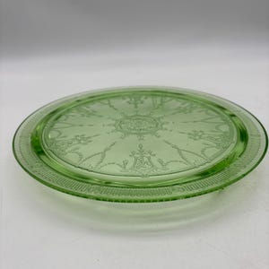 May include: A round, light green glass serving plate with a detailed, etched design. The plate has a raised rim and a central decorative pattern. The glass has a slight transparency, and the design is visible throughout the plate.