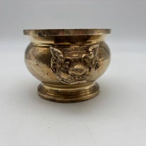 May include: A small, antique brass bowl with a detailed, embossed lion-like face on the side. The bowl has a rounded body, a flared rim, and a pedestal base. The brass has a warm, aged patina.