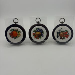 May include: Three round ceramic wall plaques with wooden frames and hanging loops. Each plaque features a different fruit design: a citrus fruit, a red apple with grapes, and a peach with berries.