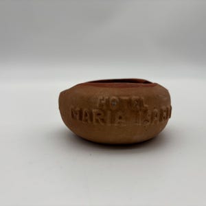 May include: A small, round, terracotta-colored bowl with the embossed text "HOTEL MARIA ISABEL" around the side. The bowl has a dark interior and sits on a white surface. The bowl is likely a souvenir or decorative item.
