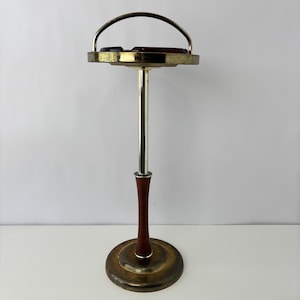 May include: A standing floor ashtray featuring a circular brass-toned tray with a handle, supported by a central metal pole with a dark brown wooden accent section, resting on a weighted, circular brass-toned base.