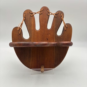 May include: A wooden wall shelf with a baseball glove design. The shelf is made of dark brown wood and features a curved shelf and finger-shaped cutouts. Leather lacing detail is visible. The shelf is supported by a small wooden peg.