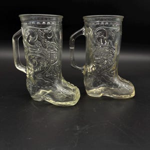 May include: Two clear glass mugs shaped like cowboy boots. Each boot-shaped mug has a handle and is decorated with an embossed floral design. The mugs are sitting on a black surface.