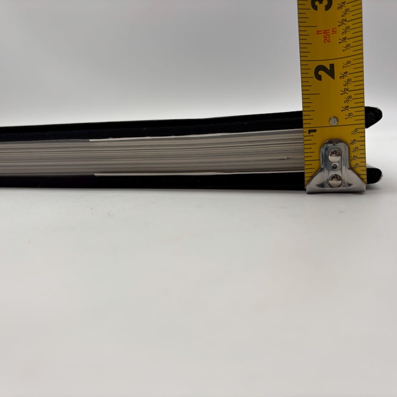 Puede incluir: Un cuaderno negro con p&aacute;ginas blancas se mide con una cinta m&eacute;trica amarilla. El cuaderno tiene aproximadamente 5 cm de grosor. La cinta m&eacute;trica est&aacute; marcada en pulgadas y fracciones de pulgada.