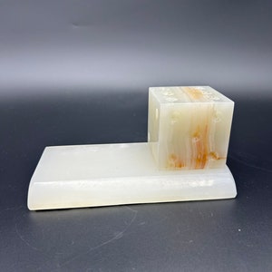 May include: A white and tan onyx desk organizer. The organizer has a rectangular base with a cube-shaped section on top. The cube has holes for pens or pencils. The onyx has natural brown and tan veining.
