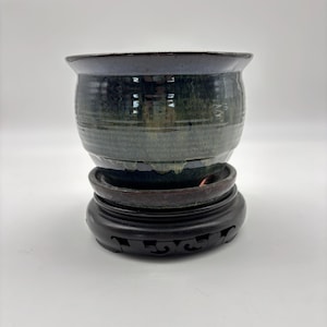 May include: A ceramic planter with a dark blue rim and a green and blue body sits on a dark brown wooden stand. The planter has a wide, open top and a rounded shape. The stand has a decorative, carved design.