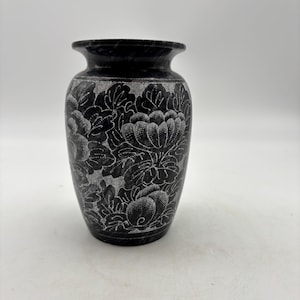 May include: A black stone vase featuring a carved floral pattern with light gray detailing. The vase has a rounded body, a flared rim, and a smooth, polished finish. The intricate engravings depict layered petals and foliage across the surface.