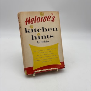 May include: A vintage cookbook titled "Heloise's Kitchen Hints" by Heloise. The cover is off-white with red and yellow accents, featuring text and floral illustrations. The book is displayed on a gold-colored wire stand, suggesting a retro kitchen theme.