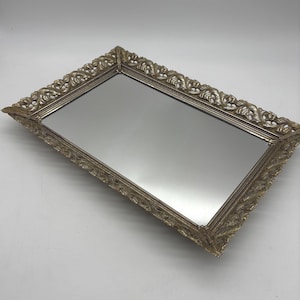 May include: A rectangular vanity tray featuring a reflective mirrored base and an ornate, metallic gold-toned frame. The border is decorated with intricate, raised scrollwork and leaf-like patterns that extend along all four edges of the tray.