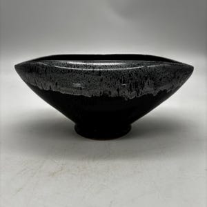 May include: A ceramic bowl with an elongated, oval-shaped rim and a pedestal base. The exterior features a glossy black finish on the lower portion, transitioning into a textured, speckled grey and white glaze along the upper rim and edges.