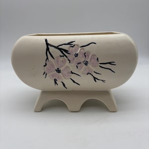 May include: Cream-colored ceramic planter with a raised floral design in lavender and black. The oval-shaped planter sits on three rounded feet. The flowers are clustered on a branch.