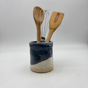 May include: A cylindrical ceramic utensil holder with a two-tone finish featuring a dark blue glazed upper section and a natural beige unglazed base. Inside the holder are a wooden spoon, a metal wire whisk, and a wooden spatula.