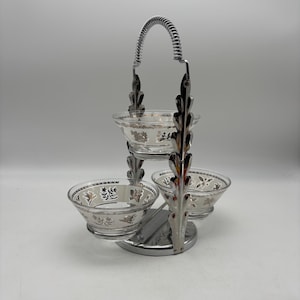May include: A three-tiered serving dish with clear glass bowls and a silver-colored metal frame. The bowls have a decorative floral pattern. The handle is a coiled design. The dish is designed for serving snacks or appetizers.