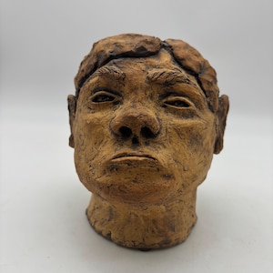 May include: A hand-sculpted clay head bust featuring a textured, earthy brown finish. The sculpture displays detailed facial features including defined eyebrows, a prominent nose, and a neutral expression, with a rough, organic surface texture.