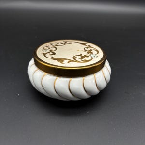 May include: A white ceramic trinket box with a gold rim and lid. The lid features a cream-colored center with gold scrollwork. The box has a ribbed design with gold accents. The box is round and sits on a black surface.