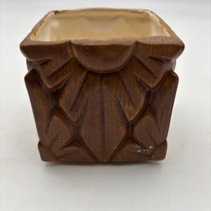 May include: A square, brown ceramic planter with a carved leaf design. The interior is a light beige color. The planter is approximately 4 inches wide and 4 inches tall, suitable for small plants or succulents.