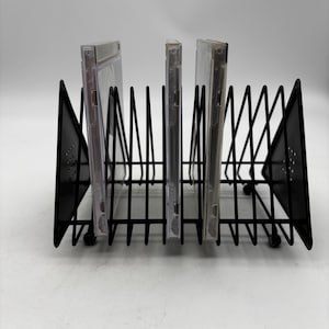 May include: A black metal wire rack designed for organizing media cases, featuring triangular side panels with small circular perforations. The rack holds three clear plastic CD cases upright in separate slots, supported by a sturdy base with small feet.