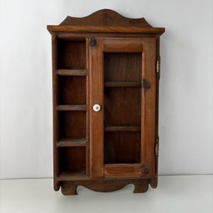 May include: A wall-mounted wooden cabinet with a medium brown finish, featuring an open shelving section with four compartments on the left and a single hinged door with a glass panel and a small white knob on the right side.
