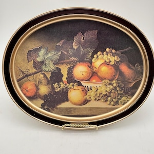 May include: Oval serving tray with a still life fruit painting. The painting depicts apples, grapes, and pears in a bowl and on a table. The tray has a dark brown border with a gold inner rim and a gold handle.