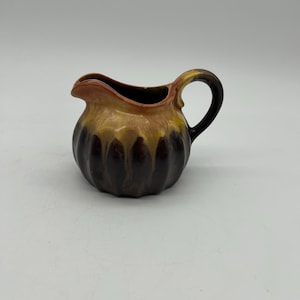 May include: A small, brown and yellow ceramic creamer with a rounded body and a handle. The creamer has a textured, ribbed design.