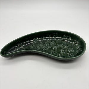 May include: A teardrop-shaped ceramic dish with a glossy dark green glaze. The interior surface features a repeating embossed pattern of small, four-petaled flower shapes. The dish has raised, curved edges and a smooth, reflective finish.
