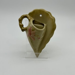 May include: A green ceramic wall vase shaped like a seashell with a pink floral design. The vase has a hole in the back for hanging.
