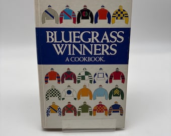 Könnte beinhalten: Ein Kochbuch mit dem Titel "Bluegrass Winners" mit weißem Einband und blauem Band mit dem Titel. Das Cover zeigt farbenfrohe Jockey-Seiden. Das Kochbuch ist ein kulinarischer Ratgeber.