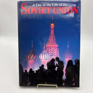 Puede incluir: Un libro de tapa dura titulado "A Day in the Life of the Soviet Union". La portada presenta una imagen nocturna de la Catedral de San Basilio en Mosc&uacute;, con siluetas en primer plano y el t&iacute;tulo en rojo.