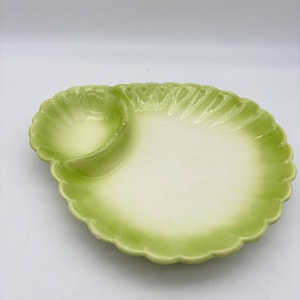 May include: A light green ceramic serving dish with a scalloped edge and a built-in dip bowl. The dish has a two-tone color scheme, with a pale green center and a darker green border. The dish is designed for serving appetizers.